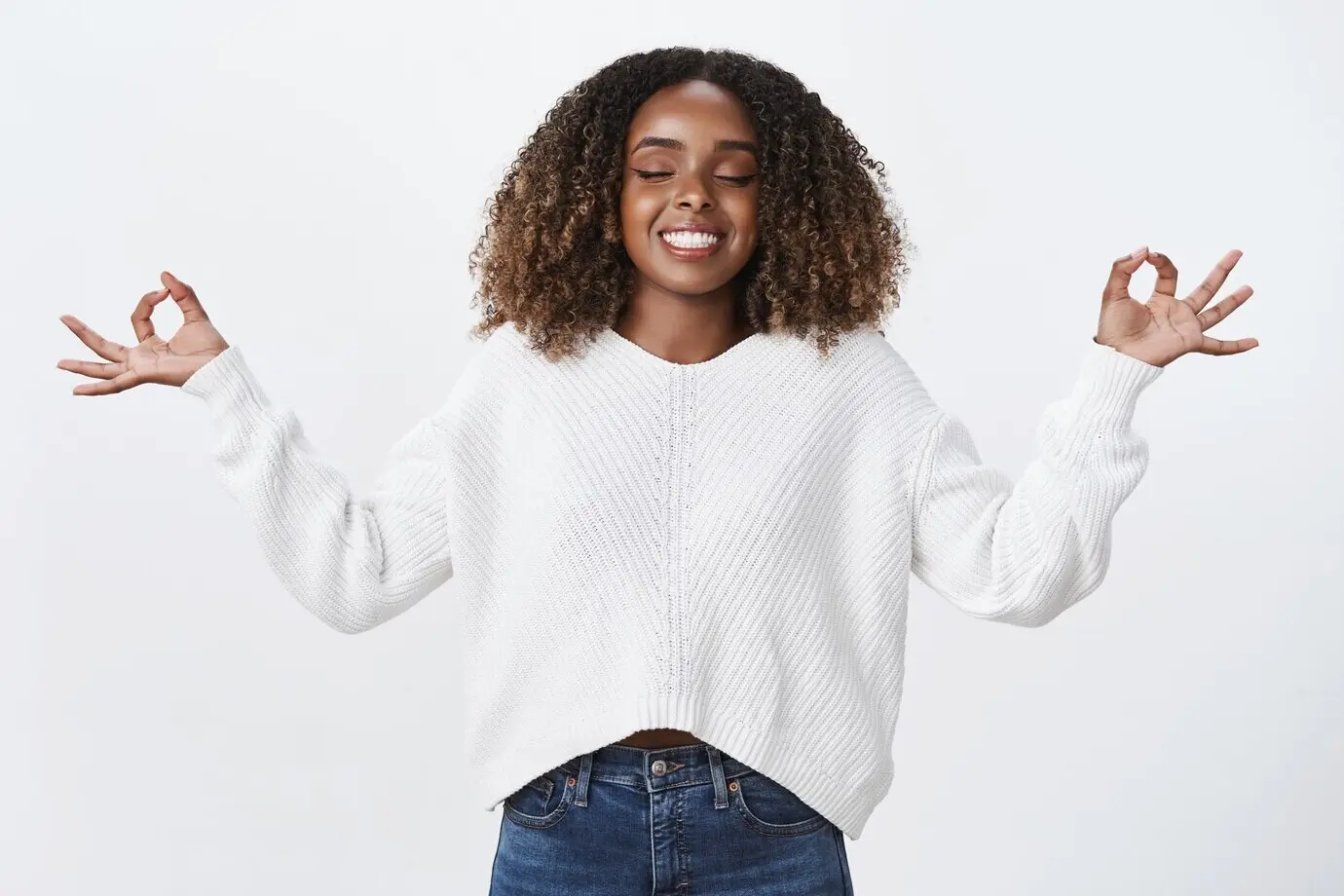 Расслабленная, испытывающая облегчение афроамериканская женщина вдыхает воздух, радостно улыбается, медитирует с закрытыми глазами, показывает «орбы», жест нирваны, в позе лотоса на фоне белой стены.