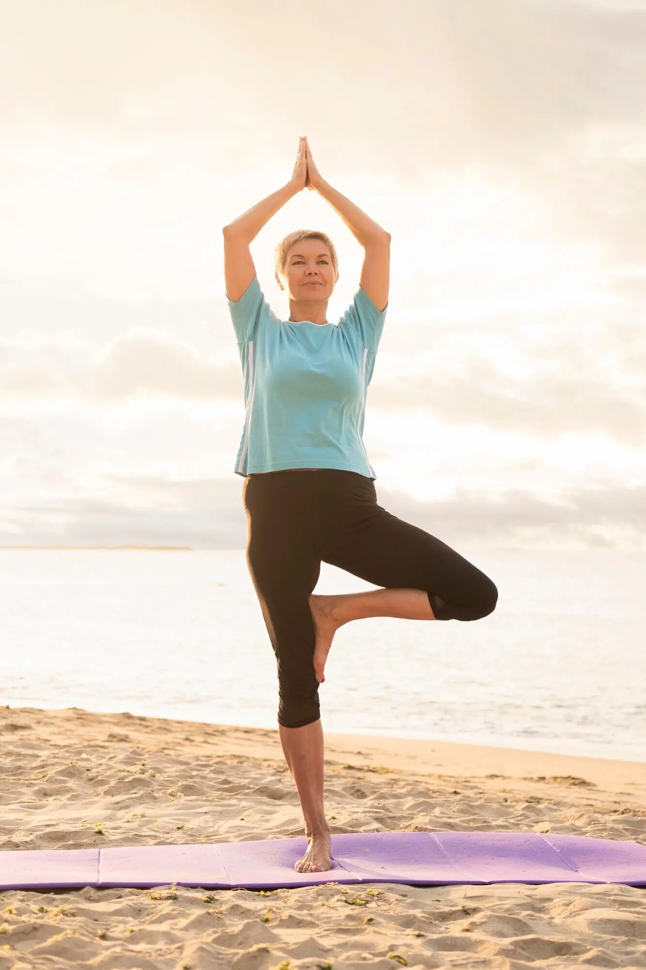 Пожилая женщина занимается йогой на пляже.